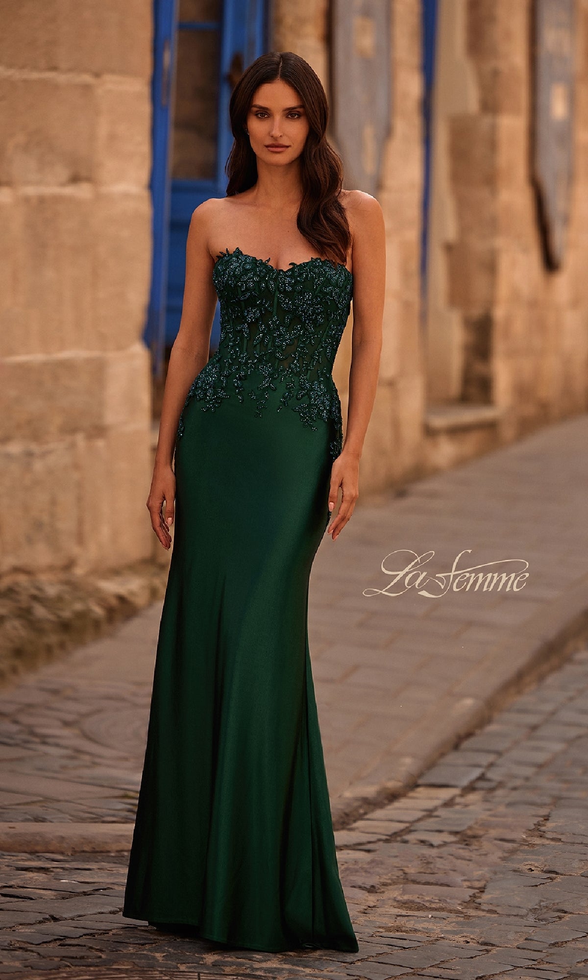 A woman stands on a cobblestone street in front of stone buildings with blue shutters, wearing the Tight Strapless Long Prom Dress: La Femme 32582, featuring a sweetheart neckline with a beaded bodice and floral embroidery.